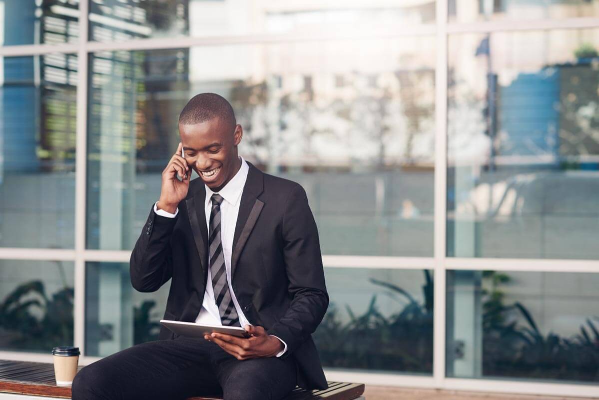 Young African businessman sitting outside his office on a bench laughing while talking on his mobile phone and looking at his digital tablet